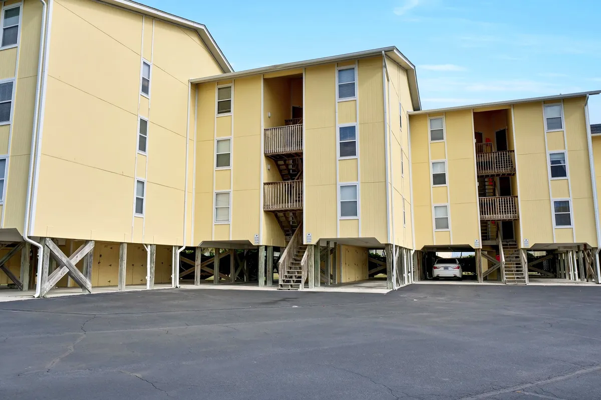 This image displays cheerful yellow buildings with visible staircases and a parking area, suggesting the exterior context and access points for guests. The outdoor staircases reflect the third-floor walk-up feature, and the welcoming environment is amplified by people relaxing on balconies and gathering with loved ones.