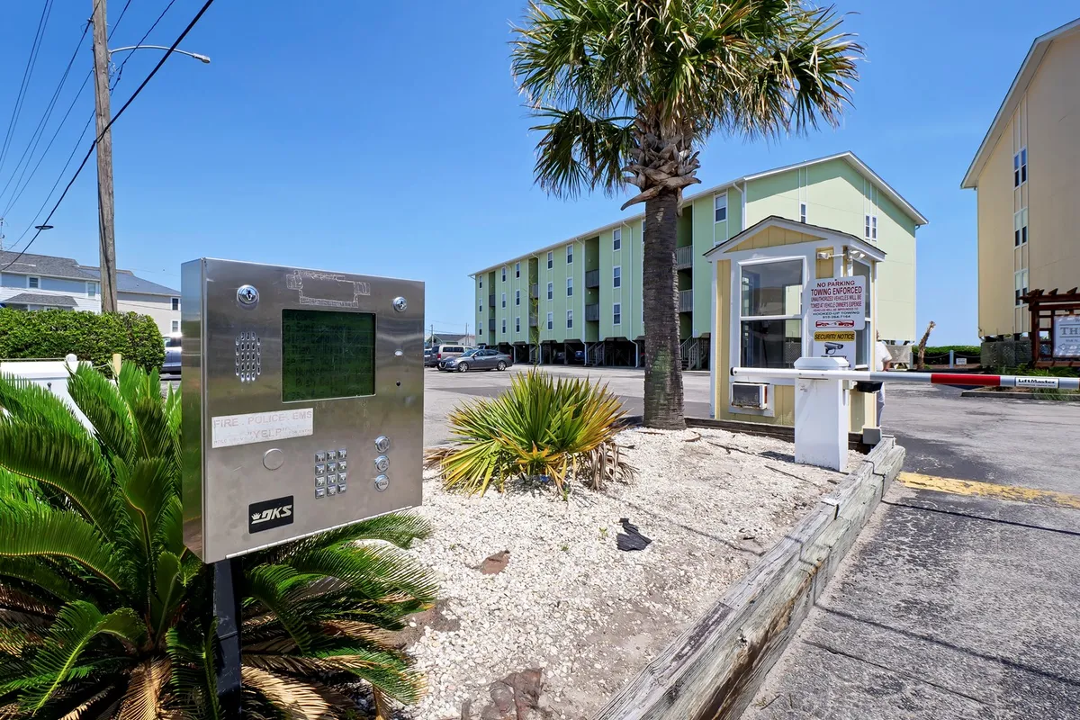 This exterior image features the building entrance framed by palm trees, an intercom system, and a welcoming parking area. The tropical landscaping and bright entry create a sense of arrival and establish a connection to the outdoor environment.