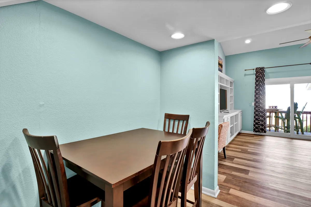 A dining table with several chairs sits beside a large window that opens to a deck. The scene is warm and inviting, suggesting a spot for family meals, game nights, or morning coffee with sunlight pouring in and a glimpse of the outdoor setting.