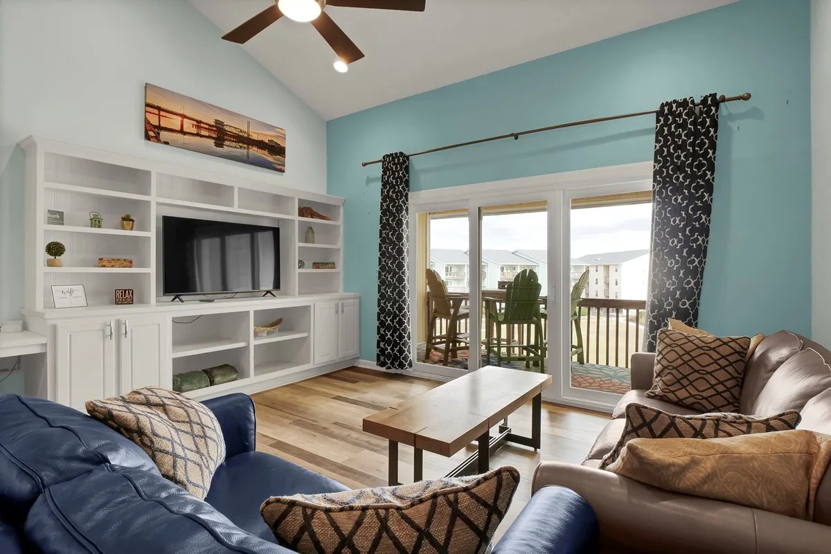 A cozy living area featuring comfortable seating, a television unit, and large windows that open out to an outdoor balcony. The room is bathed in natural light, suggesting an airy space ideal for both work and relaxation, with direct access to ocean views.