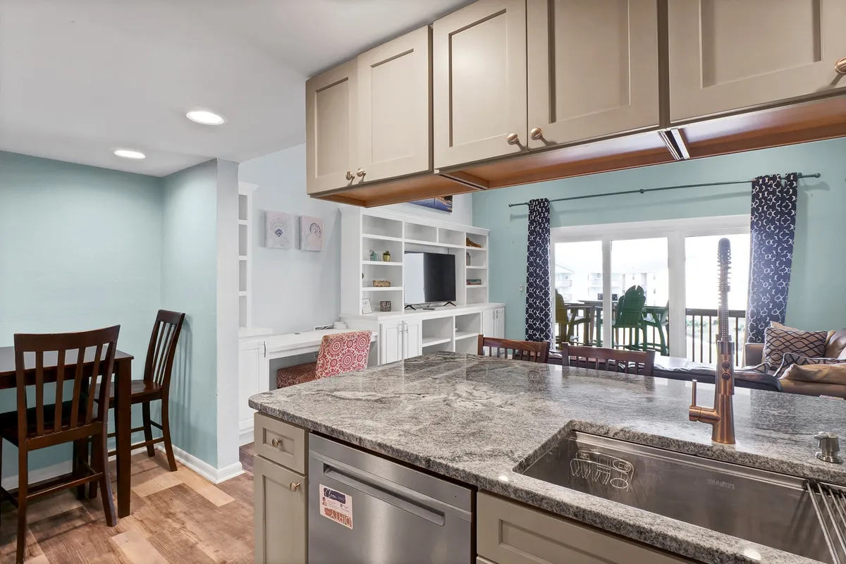 A wide shot of the communal area with a granite kitchen island, dining table, comfortable seating, and expansive windows that let in plenty of natural light. The view outside hints at a coastal atmosphere, while the layout encourages family meals and socializing.
