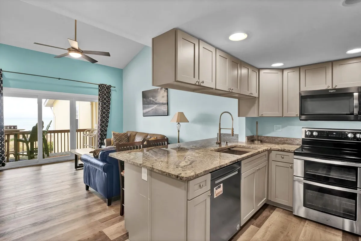 A modern, open-concept living space featuring a granite countertop, stainless steel appliances in the kitchen, a comfortable seating area, and large windows that frame ocean views. The blue accent wall adds a refreshing pop of color, while natural light streams into the area where guests can cook, relax, and dine together.
