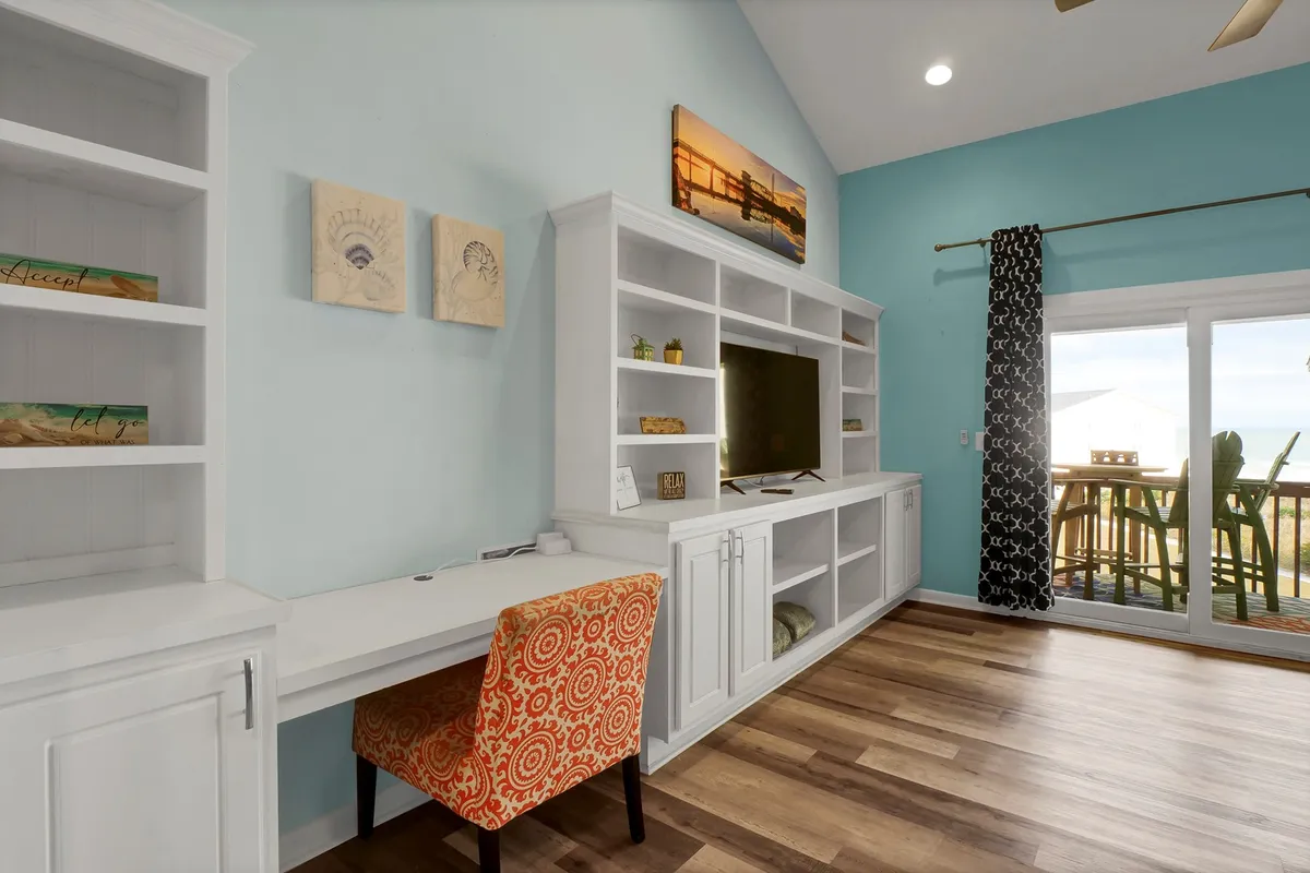 A cozy office nook with a desk and colorful chair set up next to a large TV unit and a sliding glass door. The workspace is bathed in natural light and offers a view through the sliding door, linking productivity with serenity and the beauty outside.