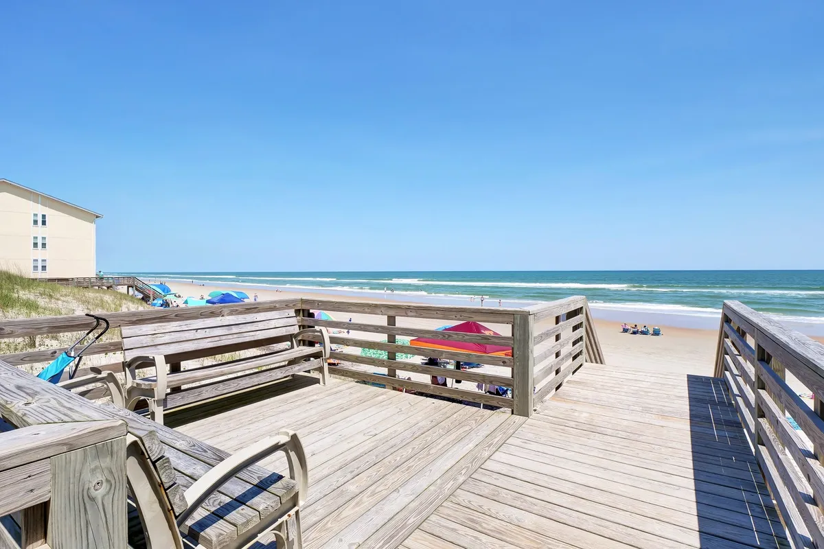 A lively wooden deck overlooking an active beach scene, with umbrellas dotting the sand, families playing, and the ocean stretching into the horizon. The deck connects the property to the vibrant beach atmosphere, emphasizing accessibility and social activity.