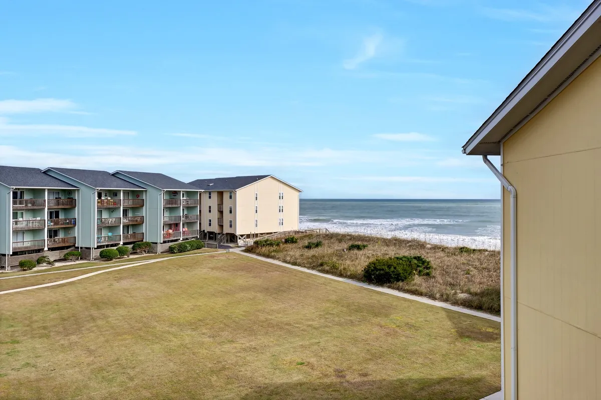 The image shows a beachfront condo complex with open green lawns leading down to sandy shores. The ocean is visible in the background, with soft sunlight suggesting either early morning or evening, and the buildings are set back just enough to hint at privacy but retain immediate beach access. The lawn invites gatherings, picnics, or games, and the entire scene feels peaceful and inviting.
