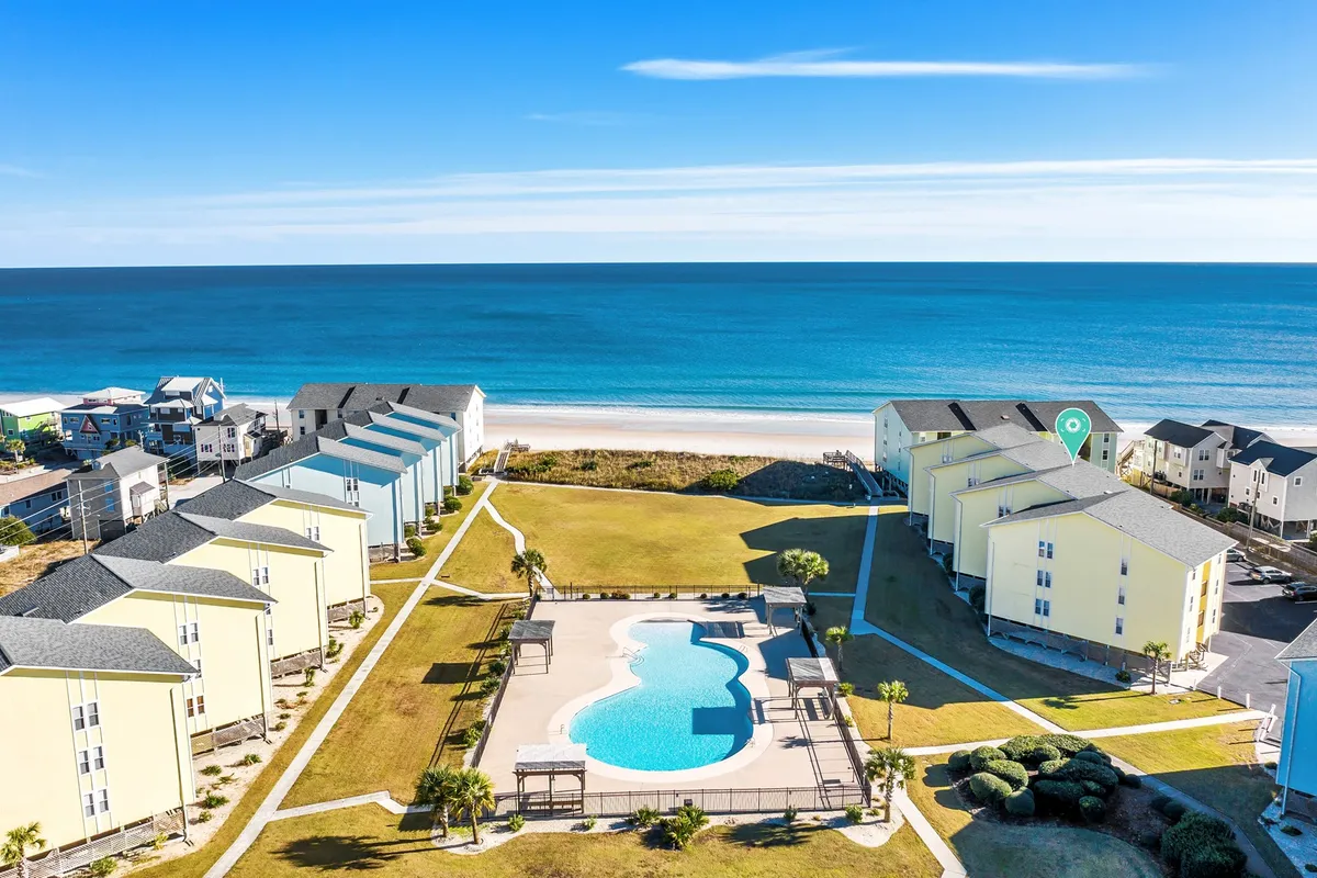 A spacious outdoor pool area with an ocean view, bordered by yellow buildings and lush landscaping. People can be seen enjoying the pool, relaxing, and making the most of the coastal lifestyle.