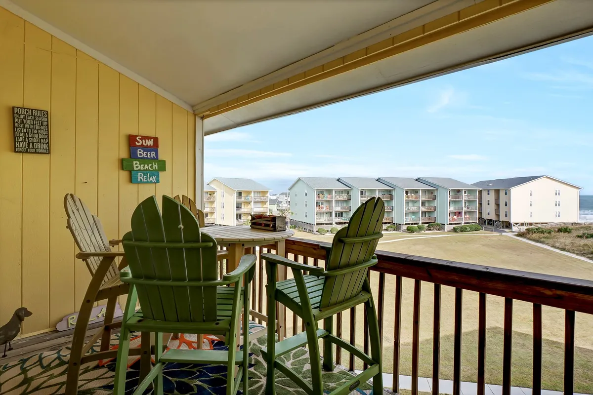 The balcony is shown with green chairs, decorative signs, and views of both the ocean and nearby buildings. The scene is lively and versatile—guests are pictured enjoying drinks, chatting, and watching the beach activity below as the sun begins to set.
